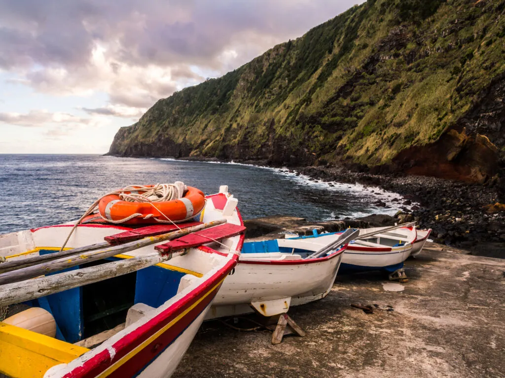 Kleurrijke houten bootjes op de kade onderaan een steile, groene rotswand langs de oceaan bij de vuurtoren van Arnel op São Miguel in de Azoren.
