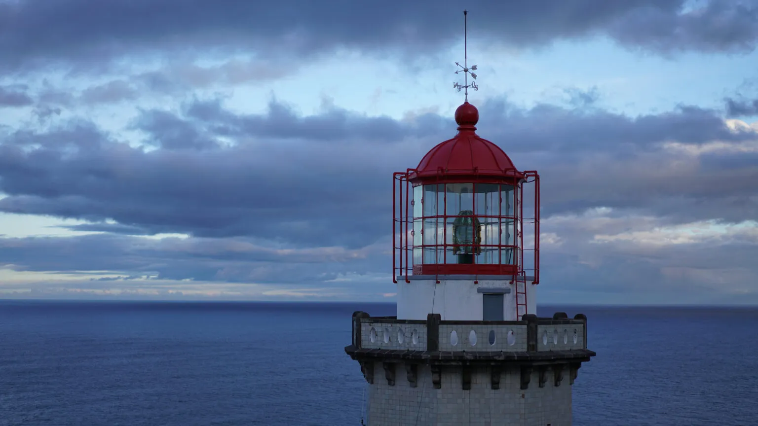 Bovenste deel van de vuurtoren Farol do Arnel met een rode koepel en lantaarnhuis tegen een schemerige lucht en een blauwe oceaan op het eiland São Miguel in de Azoren.