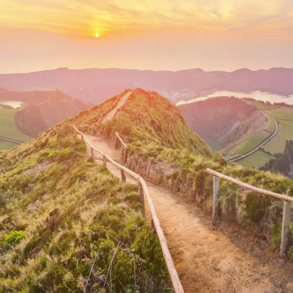 Een smal zandpad met een houten hekwerk slingert over een bergkam richting het uitzichtpunt over de kratermeren van Sete Cidades, verlicht door een warme zonsopkomst.