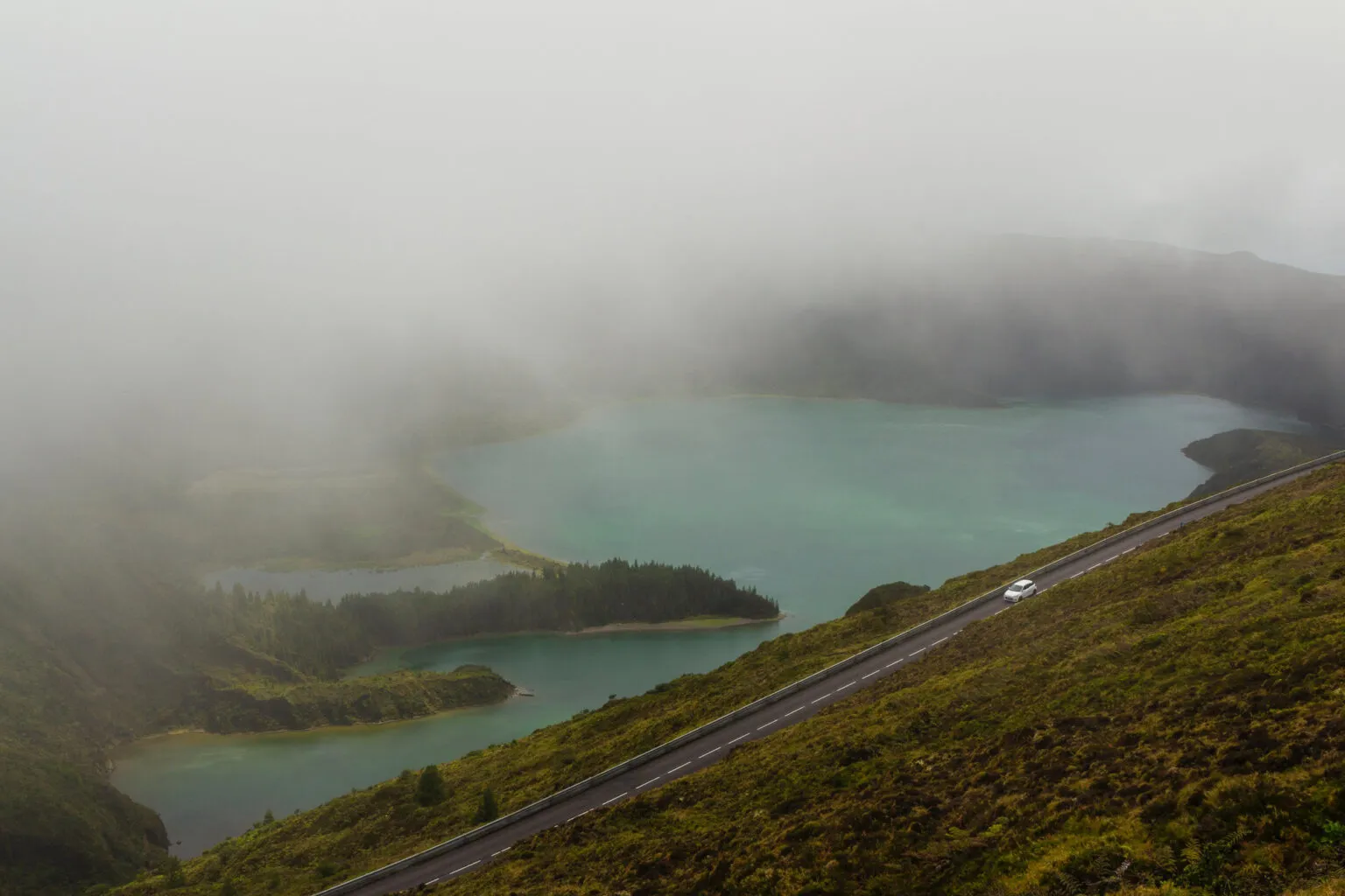 Een verharde bergweg slingert langs een turquoise kratermeer dat gedeeltelijk schuilgaat achter een dichte mistwolk.