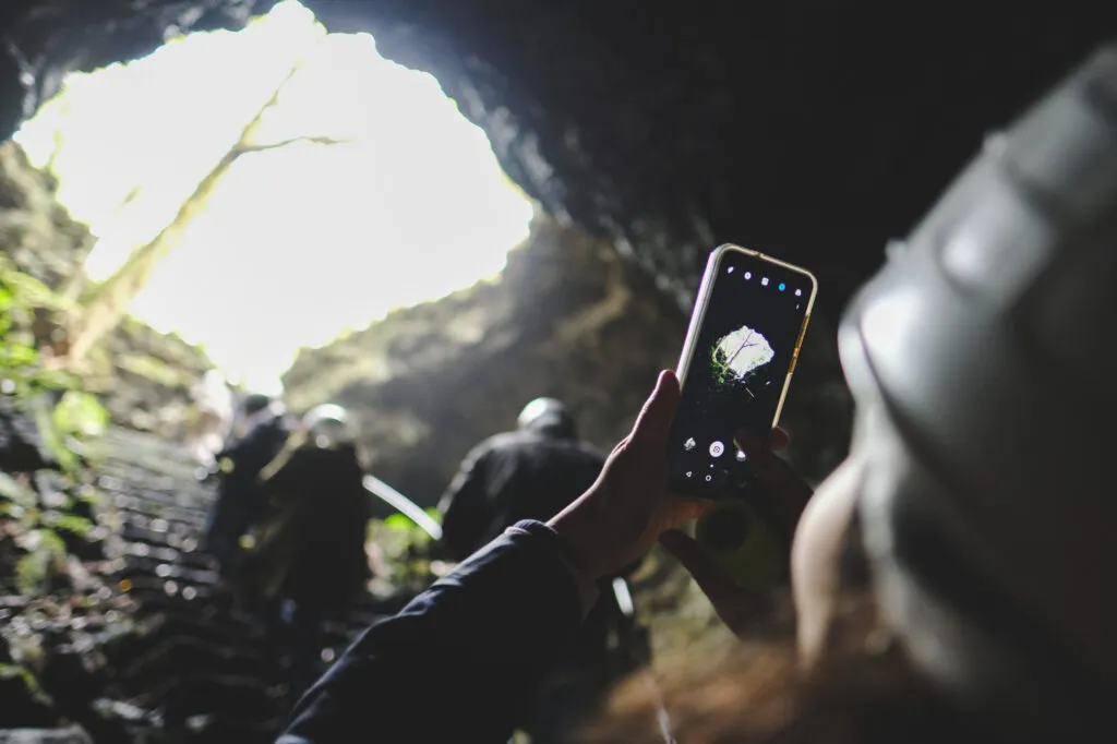 Een hand houdt een smartphone omhoog om het lichtinval bij de opening van een donkere vulkanische grot vast te leggen op de Azoren.