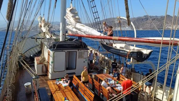 Mensen zitten aan tafels op het houten dek van een groot zeilschip op de oceaan met bergen op de achtergrond bij Tenerife.