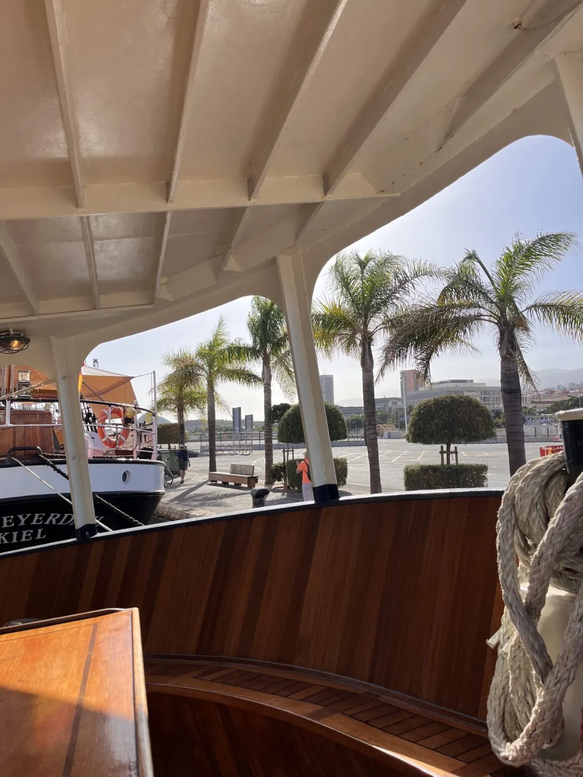 Zicht vanaf een overdekt terras met een houten balustrade op een zonnige boulevard vol palmbomen op Tenerife.