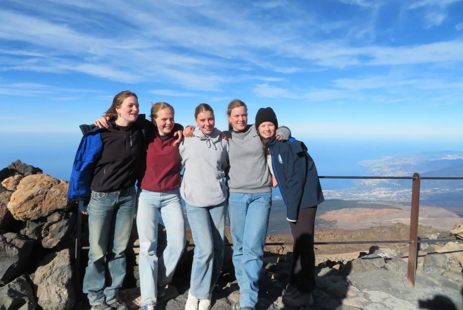 Vijf jonge vrouwen staan glimlachend arm in arm op een panoramisch uitkijkpunt bij de vulkaan Teide op Tenerife, met een helderblauwe lucht en de kustlijn op de achtergrond.