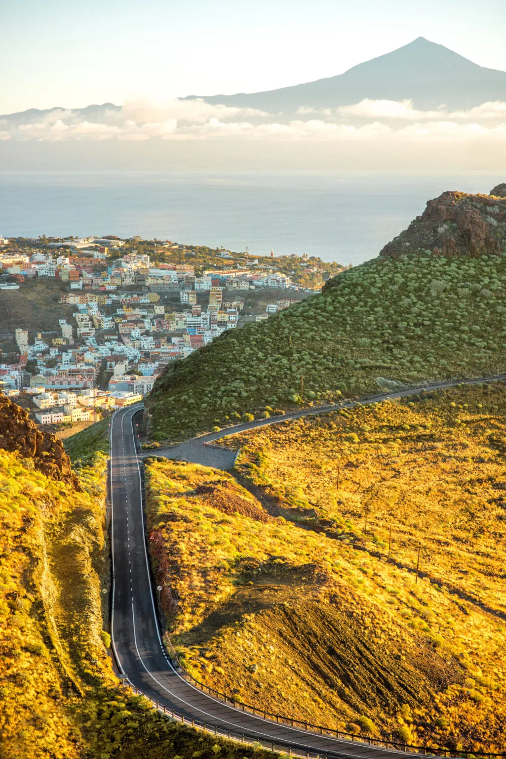 Een bergweg die afdaalt naar het kuststadje San Sebastián de La Gomera, met zicht op de oceaan en de vulkaan El Teide in de verte.