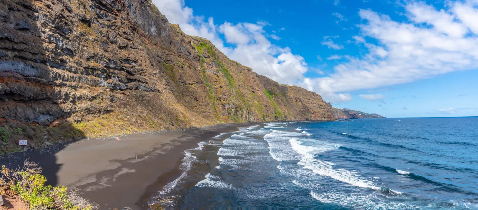 Een ruige kustlijn met het zwarte zandstrand Playa de Nogales, waar witte golfkoppen omslaan tegen de achtergrond van hoge, groene kliffen.