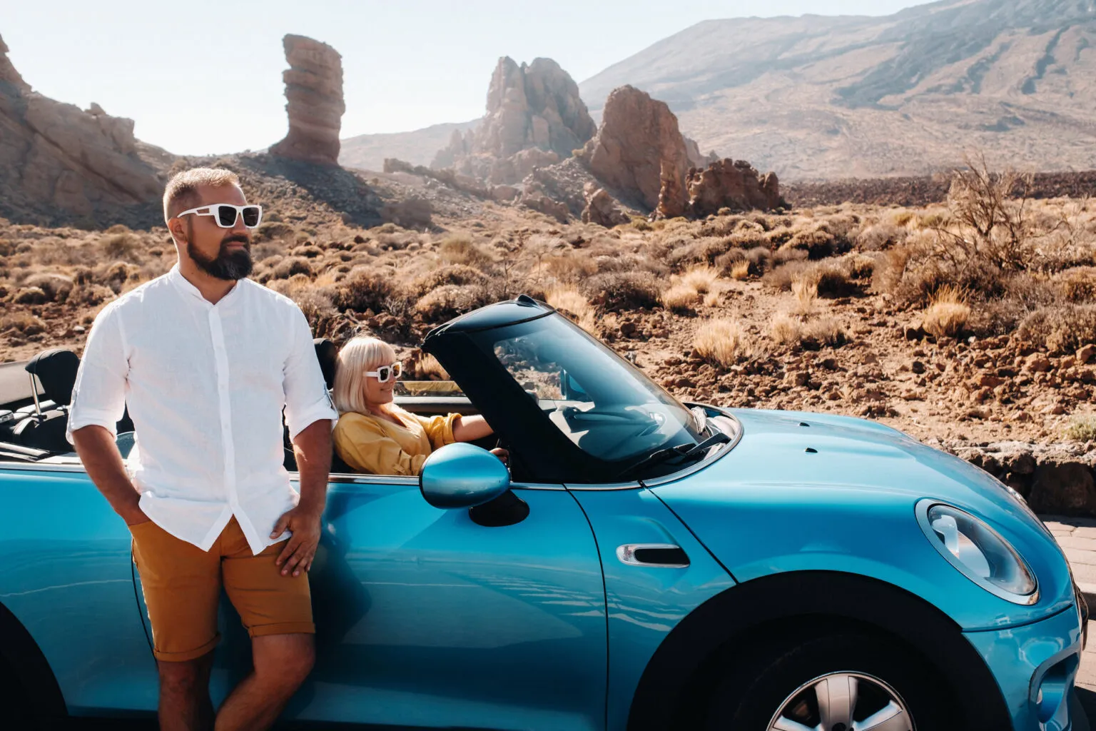 Een man leunt tegen een blauwe cabriolet waarin een vrouw achter het stuur zit, met op de achtergrond de indrukwekkende rotsformaties van El Teide op Tenerife.