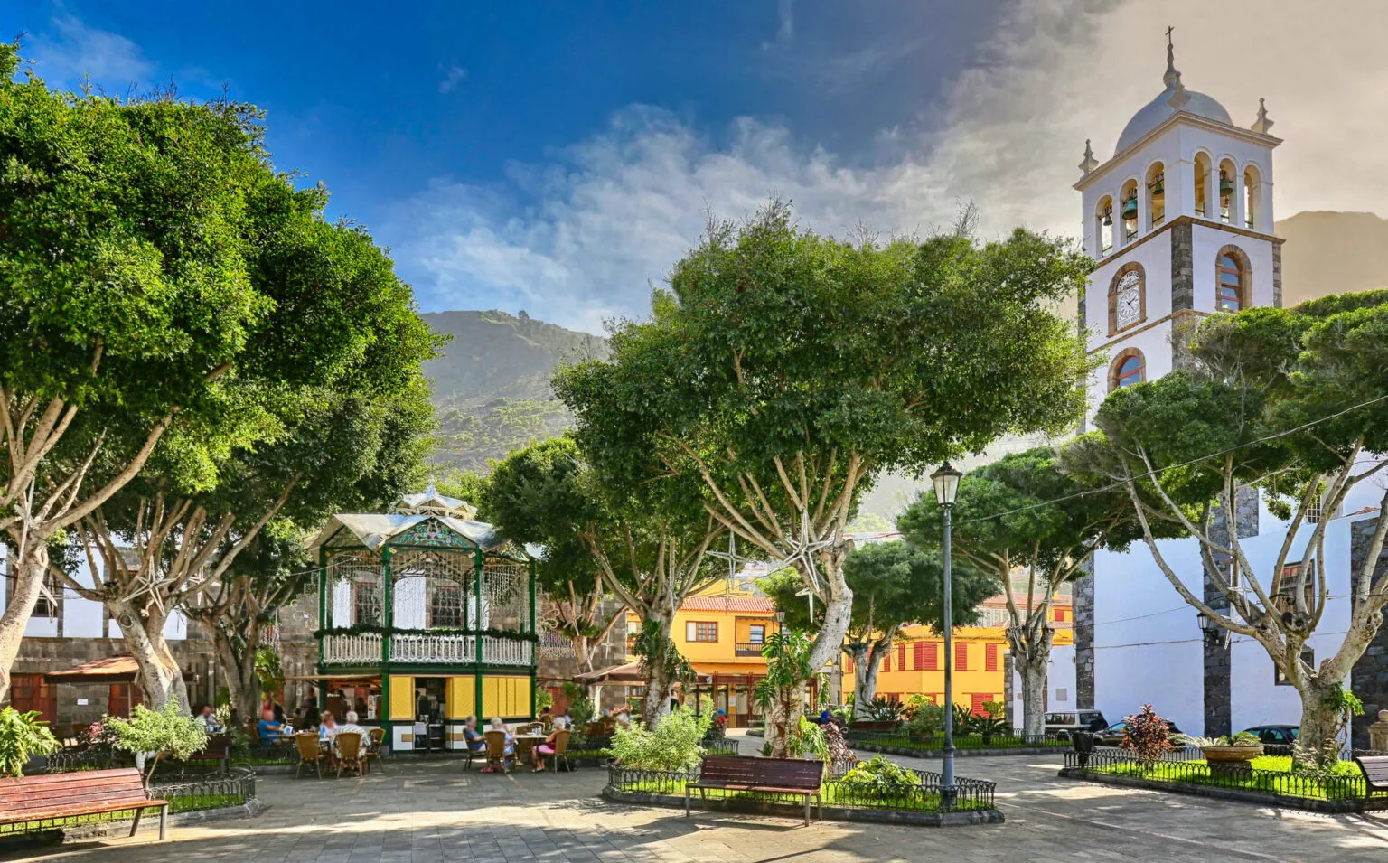 Een schaduwrijk dorpsplein met grote bomen, een gietijzeren muziektent en een witte kerktoren onder een deels bewolkte blauwe lucht in Garachico.