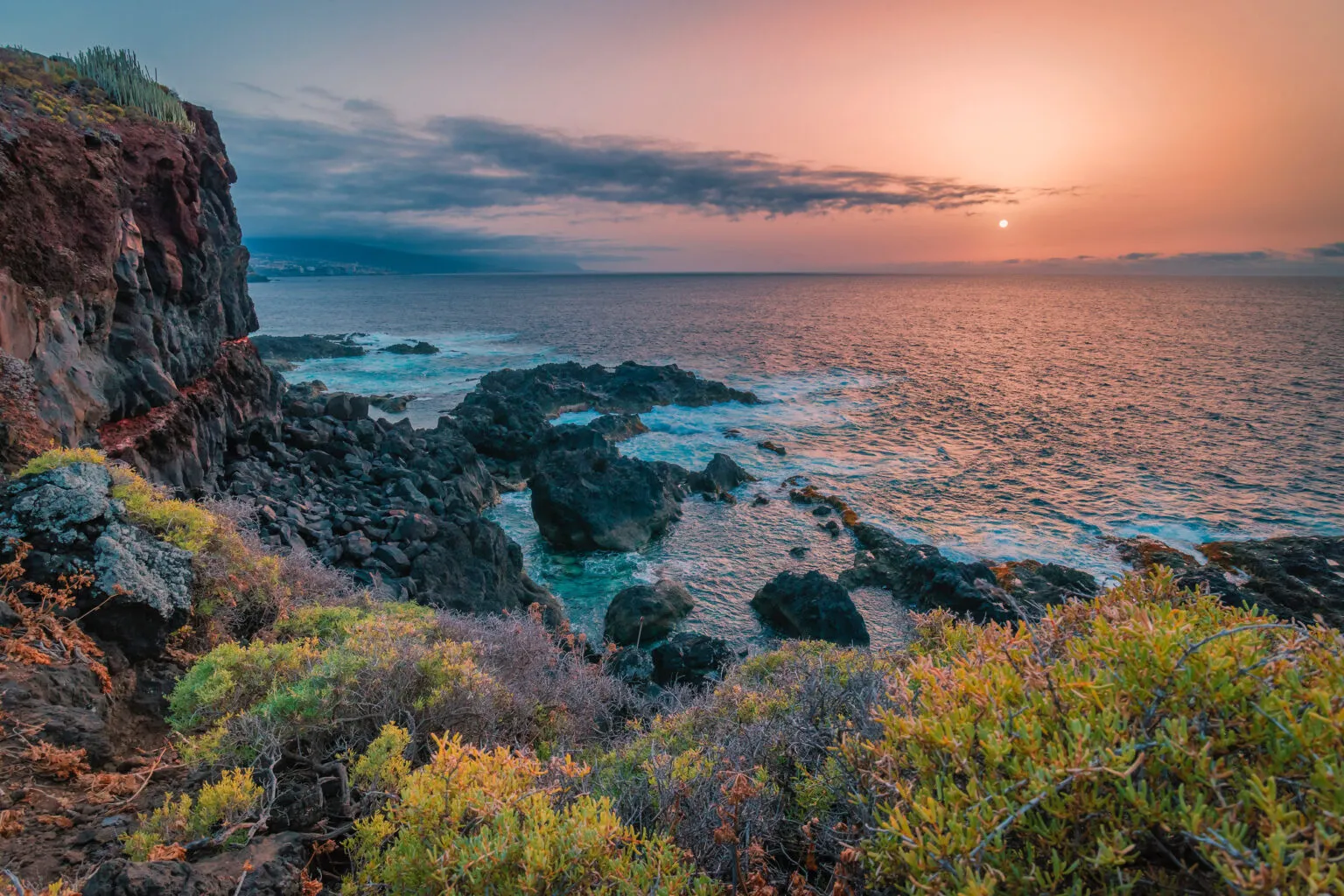 Ruige rotskust met groene begroeiing op de voorgrond en de ondergaande zon die een warme gloed werpt over de oceaan op Tenerife