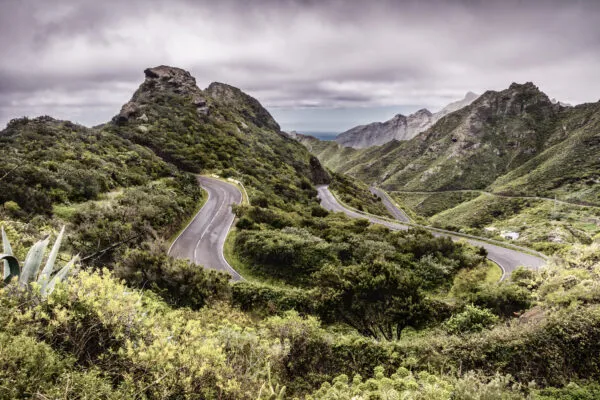 Een geasfalteerde weg meandert door de ruige, groene bergen en steile rotsformaties van het Anaga-gebergte op Tenerife onder een bewolkte hemel.