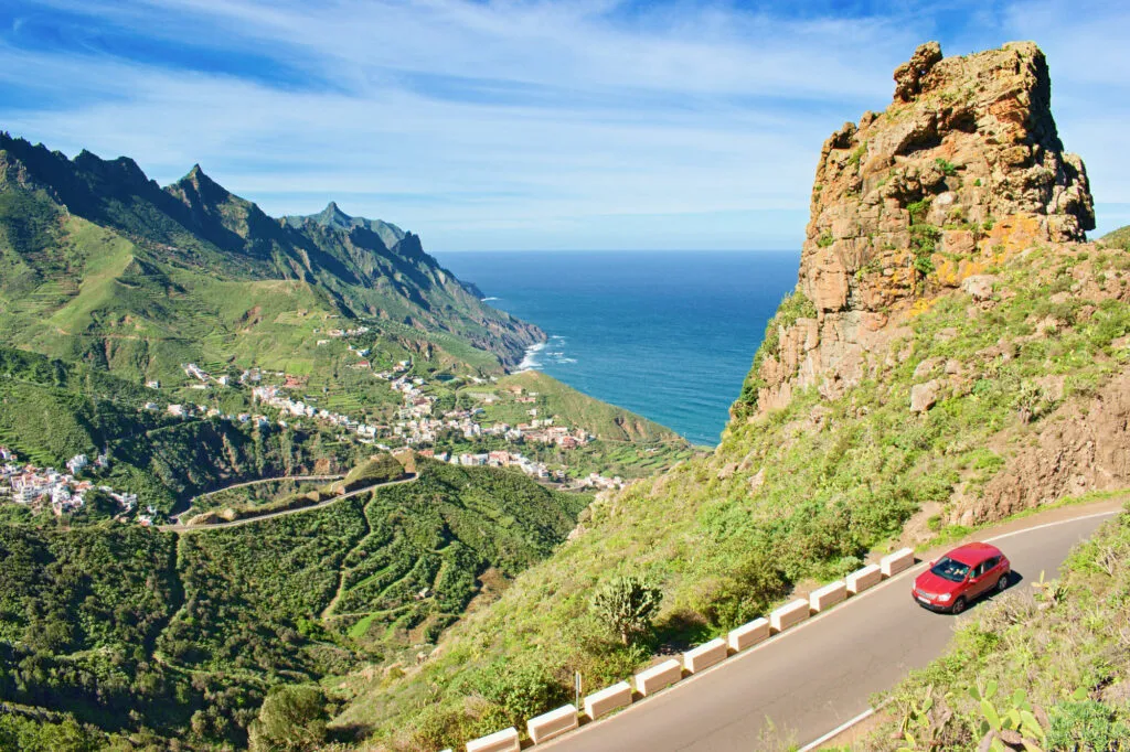 Rode auto rijdt over een kronkelende bergweg langs steile groene hellingen en een markante rots, met de oceaan op de achtergrond in het Anaga-gebergte op Tenerife
