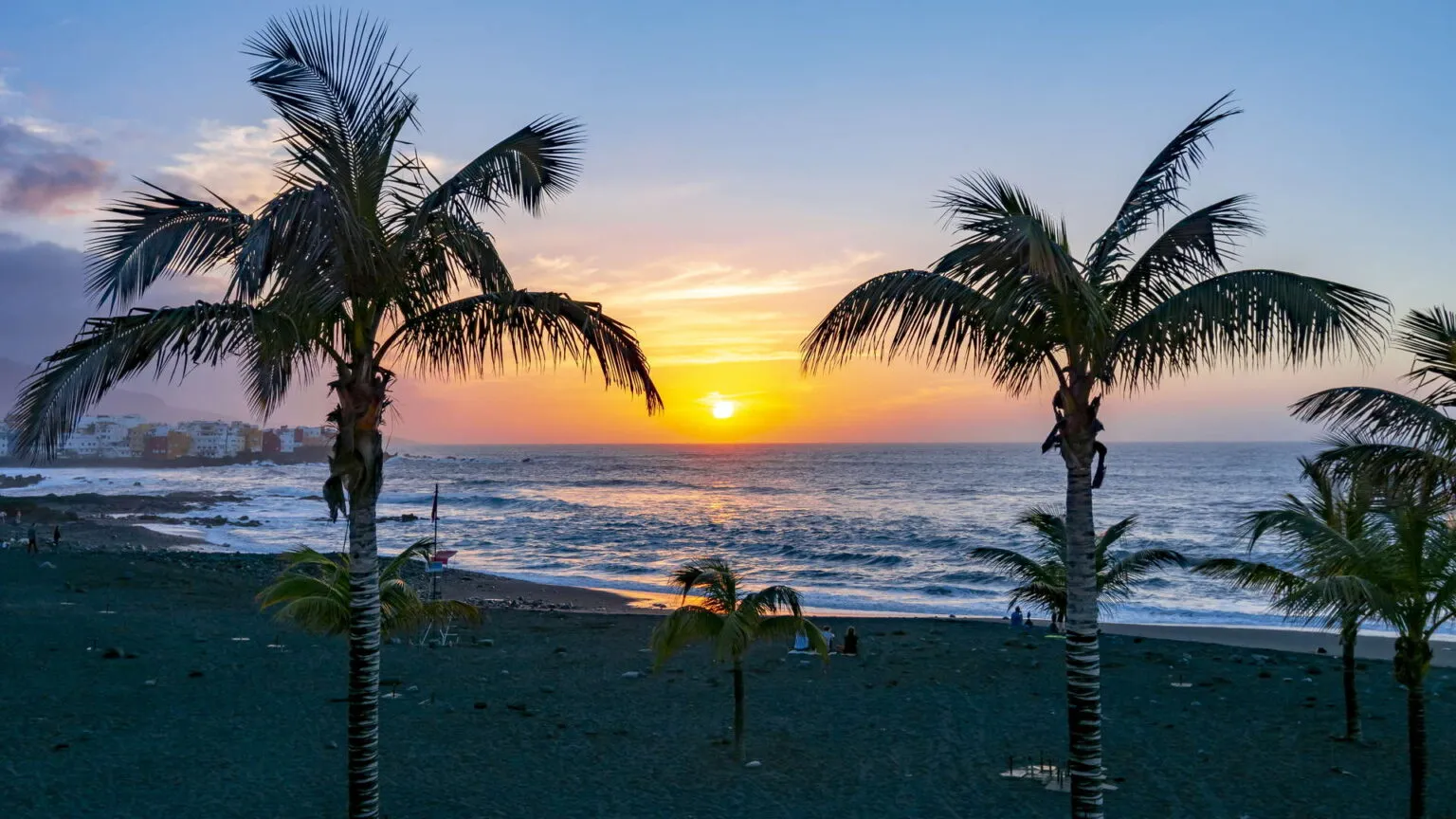 Twee hoge palmbomen in silhouet op het donkere zandstrand tegen een warme, oranje-blauwe zonsondergang boven de oceaan in Puerto de la Cruz op Tenerife