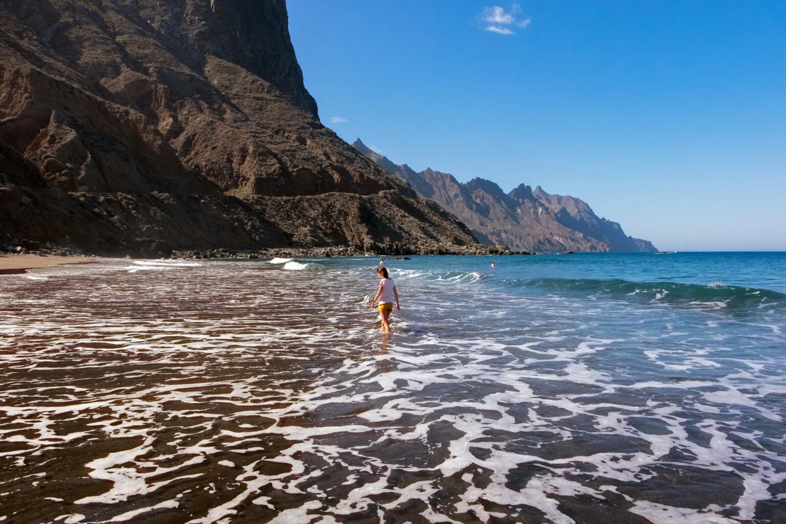 Vrouw die in het schuimende zeewater wandelt op een zwart vulkanisch zandstrand, geflankeerd door steile rotskliffen op Tenerife