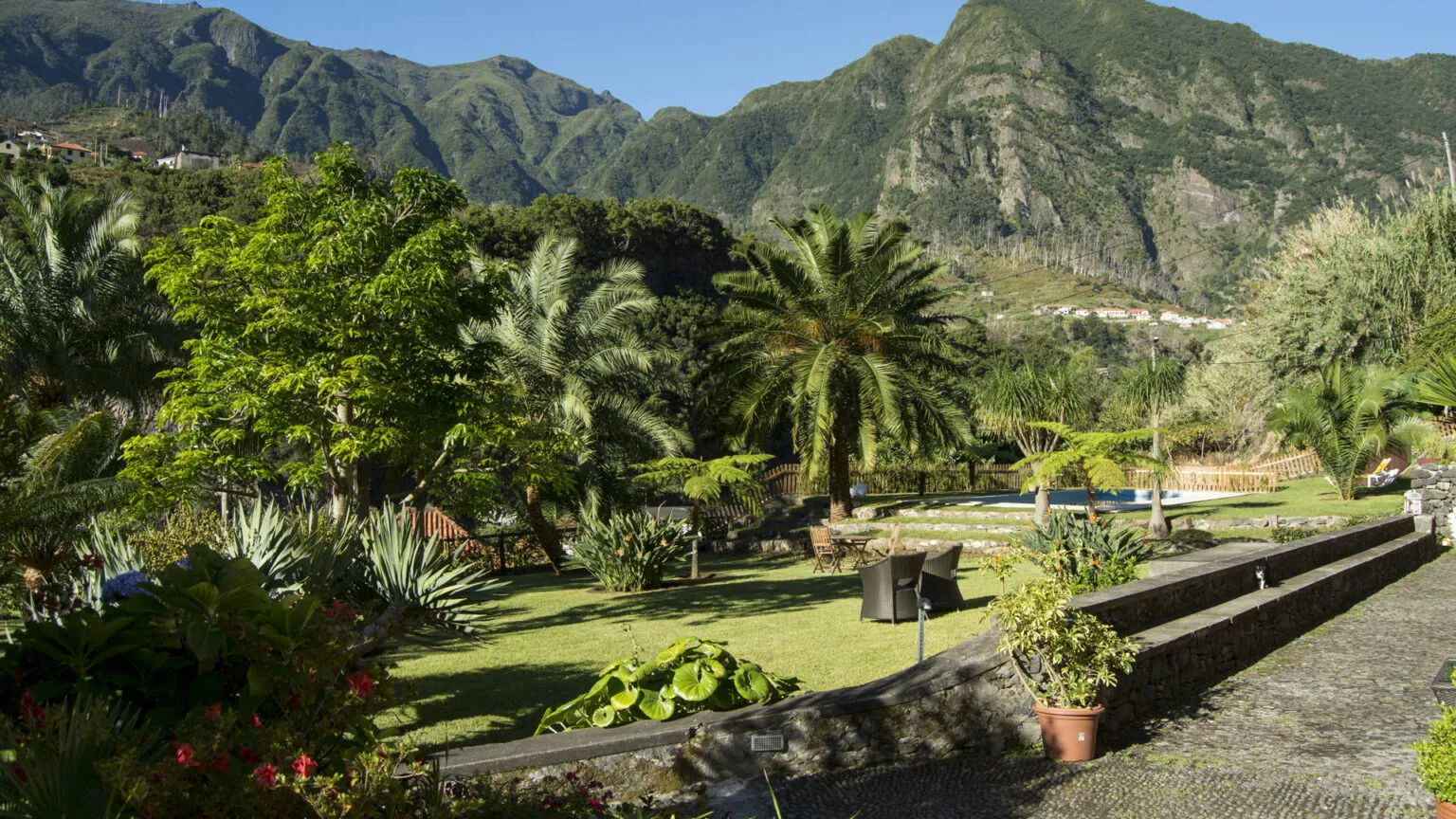 Een weelderige groene tuin met palmbomen en een stenen muurtje, met op de achtergrond een indrukwekkend uitzicht op de steile, groene bergen bij Casa da Piedade.