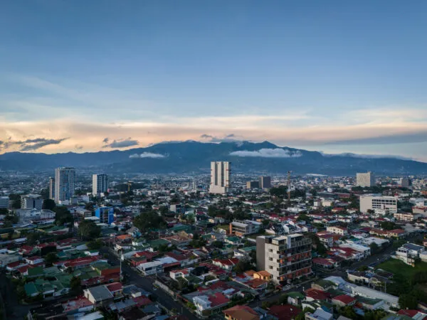 Bovenaanzicht van de uitgestrekte skyline van San José in Costa Rica, met talloze gekleurde daken en hoge gebouwen tegen een achtergrond van imposante bergen onder een opklarende avondlucht.