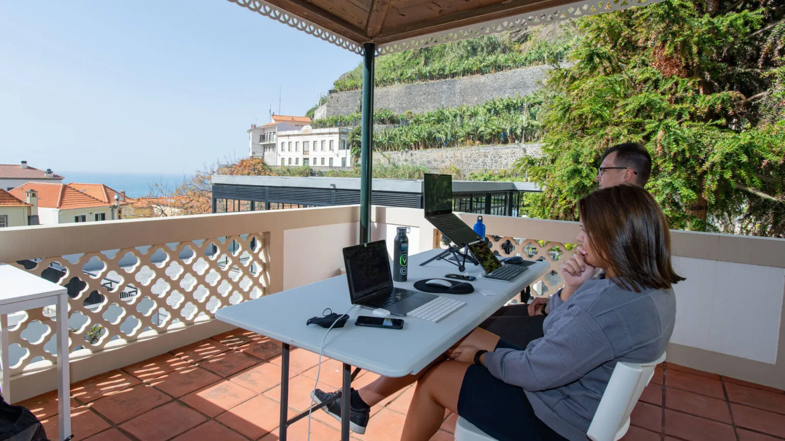 Een digital nomad zit aan een tafel met een laptop te werken op een overdekt balkon met uitzicht op de oceaan en groene heuvels op Madeira.