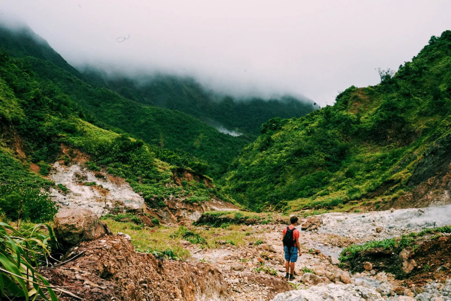 Een hiker met een rugzak staat op een rotsachtig pad in een vulkanisch dal en kijkt uit naar de mistige, dichtbegroeide groene bergen in het binnenland van Dominica.
