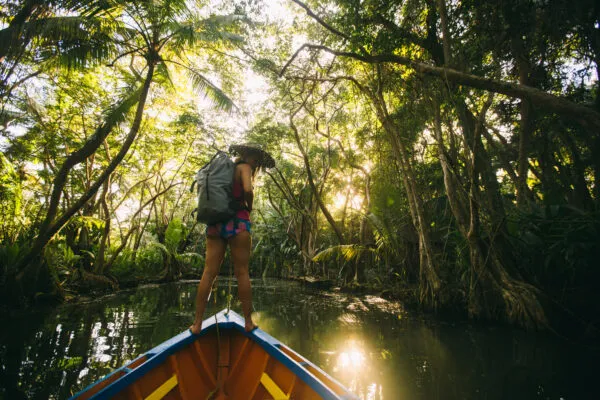 Een vrouwelijke reiziger met rugzak staat op de punt van een houten boot tijdens een boottocht over de rustige Indian River op Dominica, omgeven door dichtbegroeide junglebomen en gefilterd zonlicht.