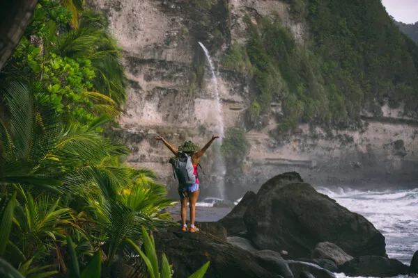 Een vrolijke vrouwelijke reiziger met een rugzak staat met de armen omhoog op een rotsachtig uitzichtpunt bij Secret Beach op Dominica.