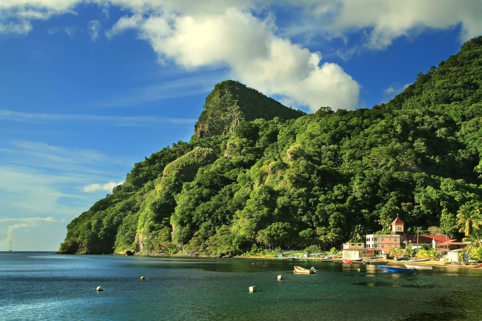Pittoresc zicht op het dorp Soufrière aan het rustige zeewater op Dominica, gedomineerd door een steile, dichtbegroeide groene bergwand onder een helderblauwe lucht.