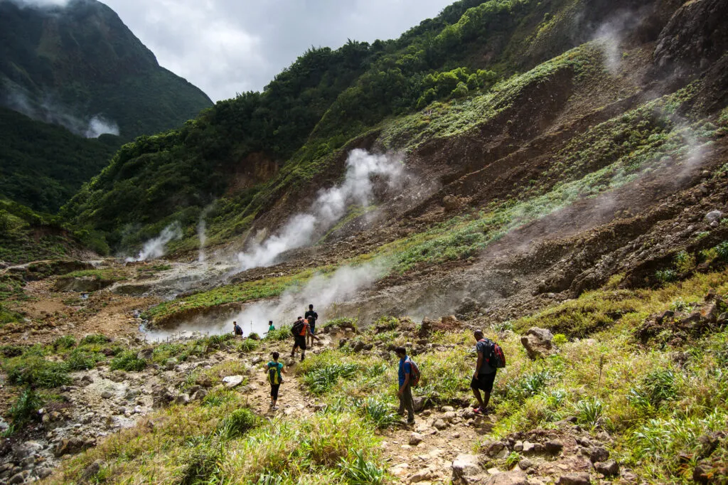 Een groep reizigers wandelt over een rotsachtig pad door de Valley of Desolation op Dominica, met stoom uit vulkanische bronnen en een steile, groenbegroeide berghelling op de achtergrond.