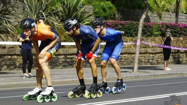 Drie inlineskaters met helmen en sportkleding rijden in volle vaart achter elkaar over de weg tijdens een skatewedstrijd.