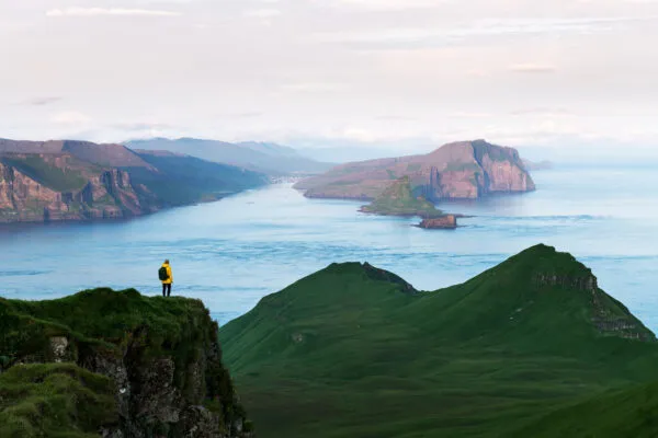 Een reiziger staat op een groene bergtop uit te kijken over de indrukwekkende fjorden en bergen rond Funningur op het eiland Eysturoy van de Faeröer Eilanden.