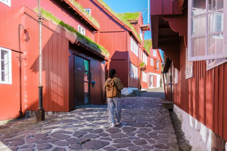 Een vrouwelijke reiziger wandelt tussen traditionele rode gebouwen met grasdak in de stad Tórshavn