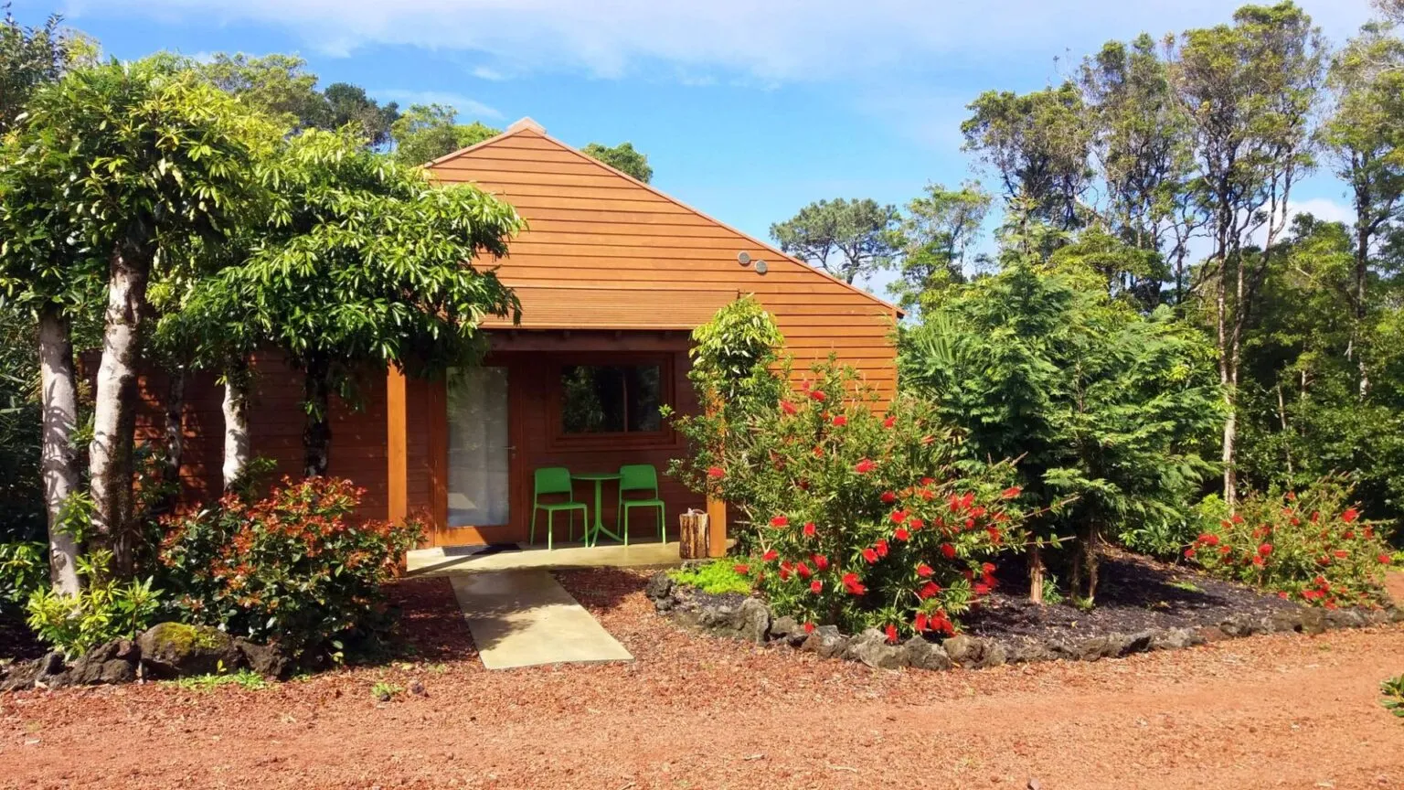 Vooraanzicht van een houten bungalow van Alma do Pico Nature Residence omgeven door groene struiken en rode bloemen op het eiland Pico.