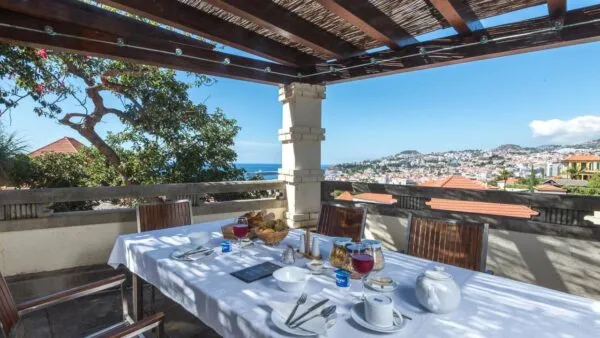 Een met een wit laken gedekte ontbijttafel op het terras van Arts Hotel Conde Carvalhal met uitzicht over de stad Funchal en de oceaan op Madeira.