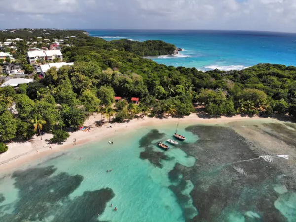 Luchtfoto van het zandstrand van Plage de Petit-Havre op Grande-Terre, Guadeloupe, met dobberende bootjes in het heldere, turquoise water nabij een dicht begroeide kustlijn met bomen.