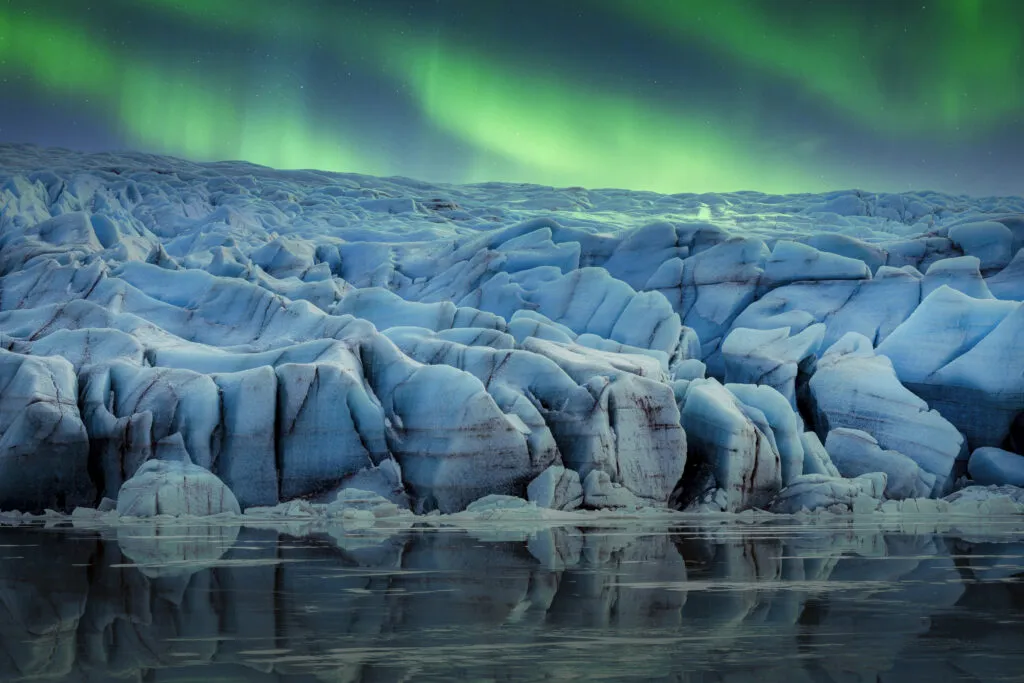 Felle groene bogen van het noorderlicht dansen aan de nachthemel boven de massieve, diepblauwe ijsstructuren van een gletsjer in IJsland, met een subtiele weerspiegeling in het voorliggende water.