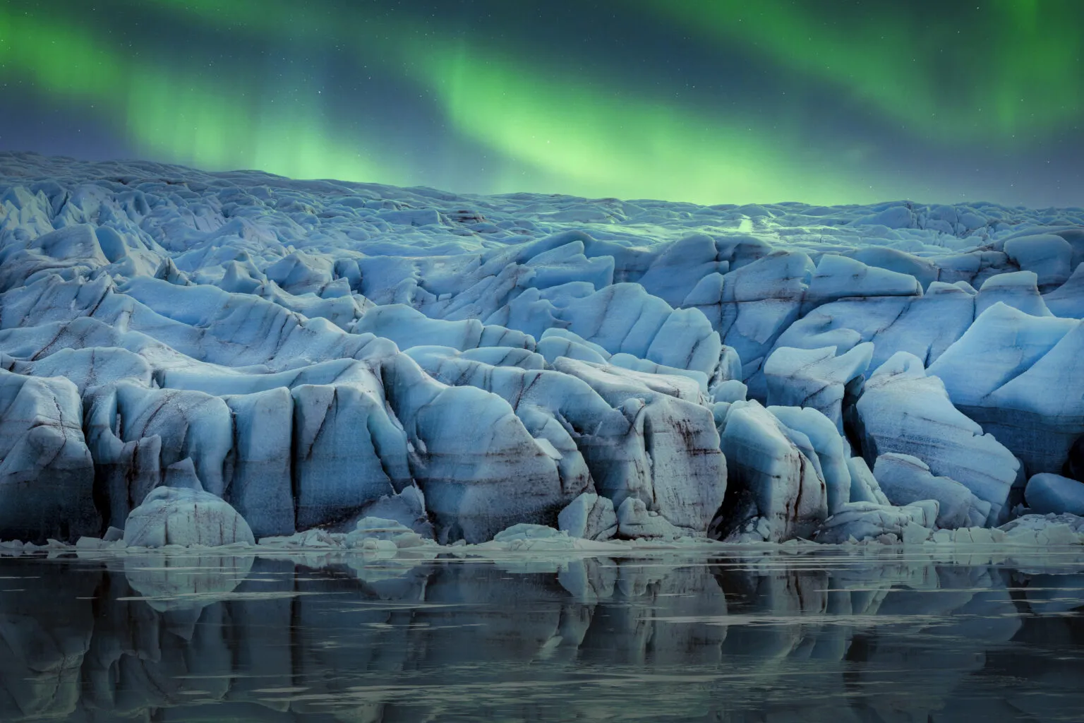 Felle groene bogen van het noorderlicht dansen aan de nachthemel boven de massieve, diepblauwe ijsstructuren van een gletsjer in IJsland, met een subtiele weerspiegeling in het voorliggende water.