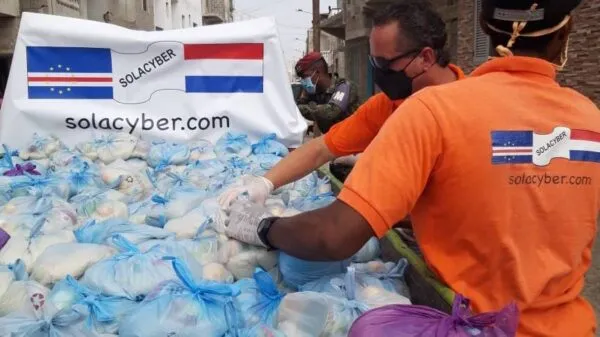Hulpverleners in oranje shirts sorteren een grote hoeveelheid plastic zakken met voedselpakketten voor een spandoek van Solacyber met de Kaapverdische en Nederlandse vlag.
