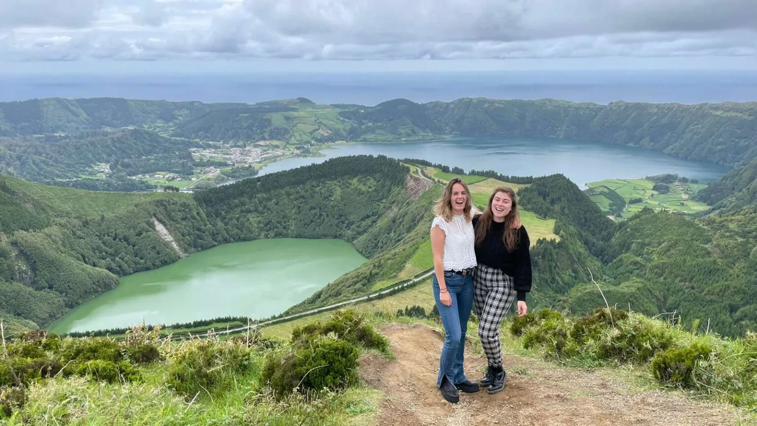 Twee vriendinnen staan arm in arm op een wandelpad, met op de achtergrond het panoramische uitzicht op de groene en blauwe kratermeren van Sete Cidades op het eiland São Miguel.