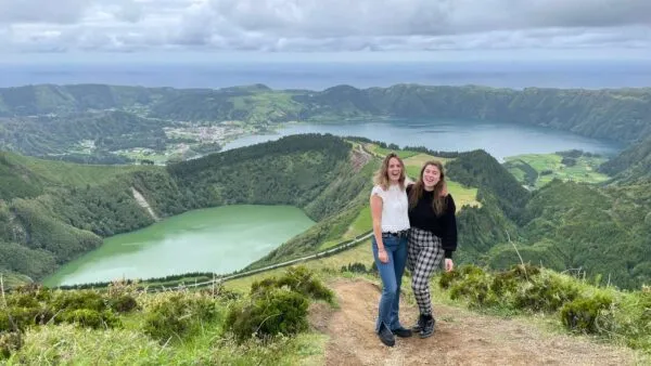 Twee vriendinnen staan arm in arm op een wandelpad, met op de achtergrond het panoramische uitzicht op de groene en blauwe kratermeren van Sete Cidades op het eiland São Miguel.
