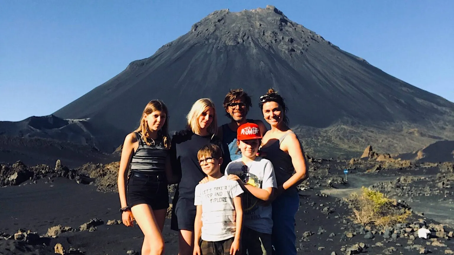 Een groep van zes reizigers poseert samen op het zwarte vulkanische landschap met de vulkaan Pico do Fogo op de achtergrond, Kaapverdië.