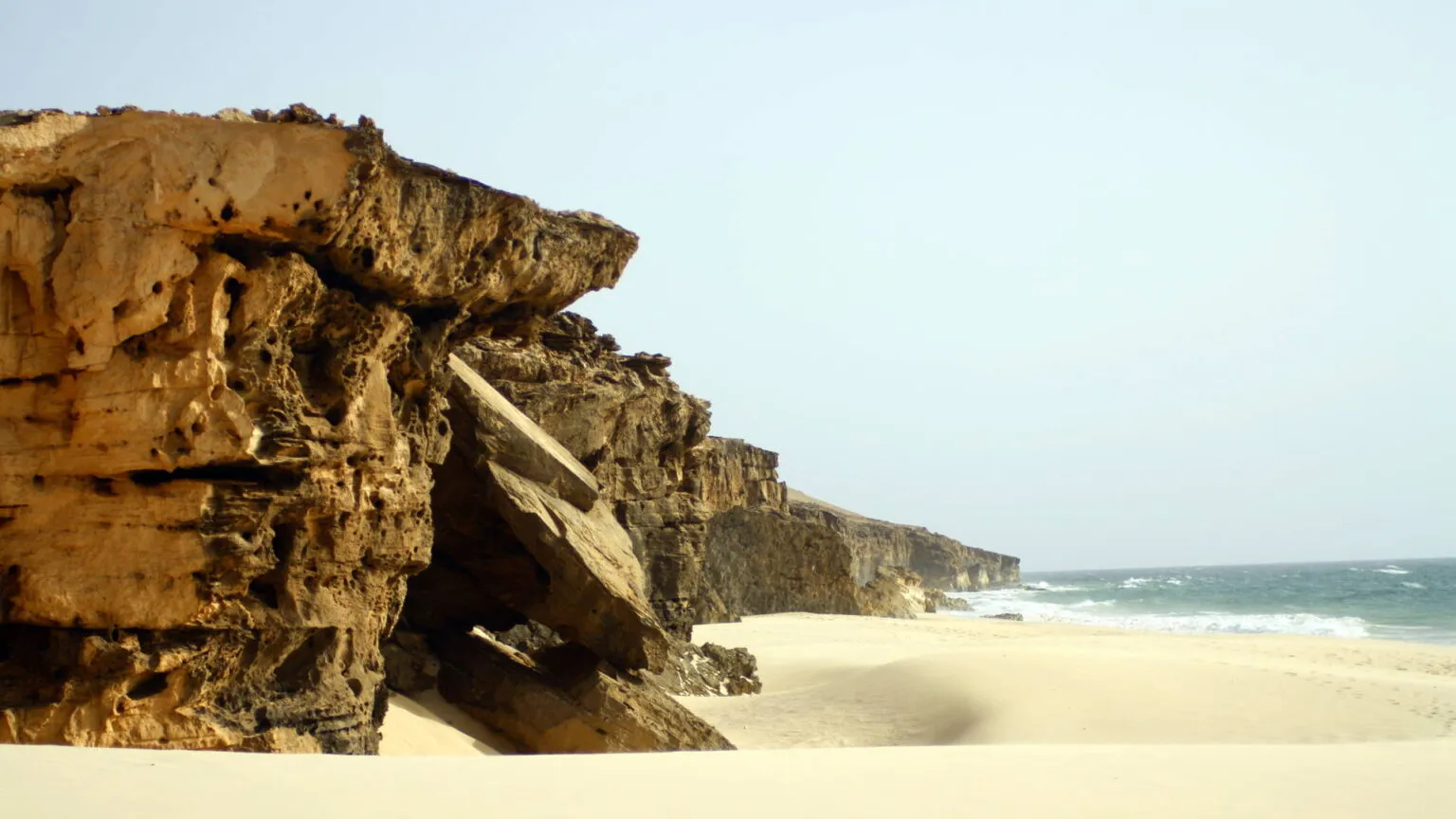 Uitgestrekt zandstrand langs een ruige, donkere rotswand en een heldere zee op Boa Vista, Kaapverdië.