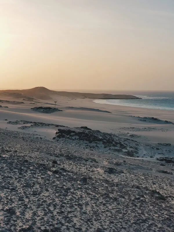 Een weidse baai met een goudgeel zandstrand en duinen die overgaan in het turquoise oceaanwater bij zonsondergang op het eiland Boa Vista, Kaapverdië.