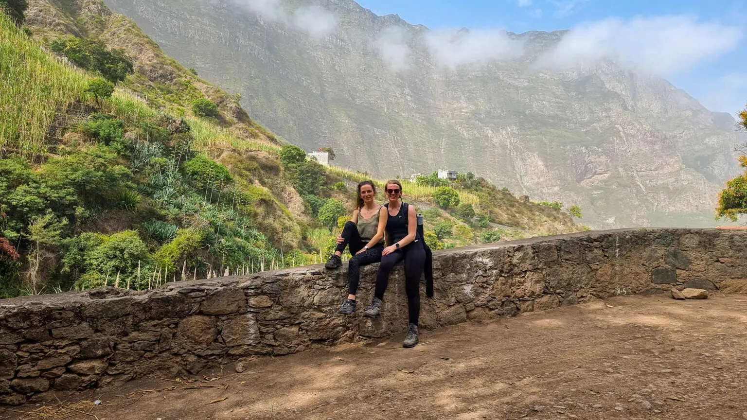 Twee wandelaars zitten op een lage stenen muur en kijken uit over het weelderige groene berglandschap van Santo Antao in Kaapverdië.