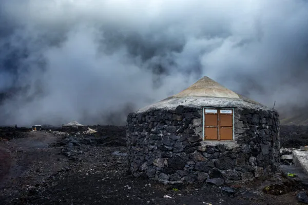 Een rond huisje opgebouwd uit zwarte lavastenen met een conisch dak in een mistig vulkanisch landschap in Chã das Caldeiras op Fogo, Kaapverdië.