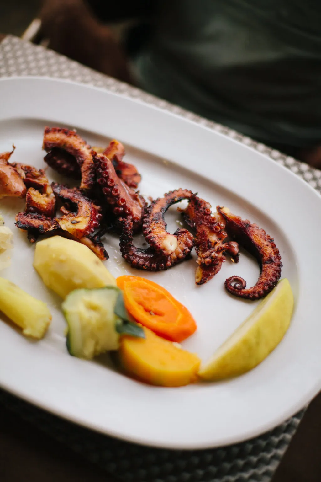 Een bord met gegrilde tentakels van octopus, geserveerd met gekookte aardappelen, wortel en een schijfje citroen.
