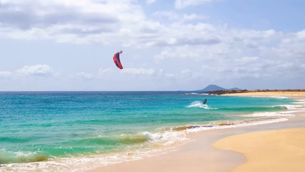 Een kitesurfer glijdt met een rode kite over de helderblauwe oceaan langs het zandstrand van Santa Maria op Sal.