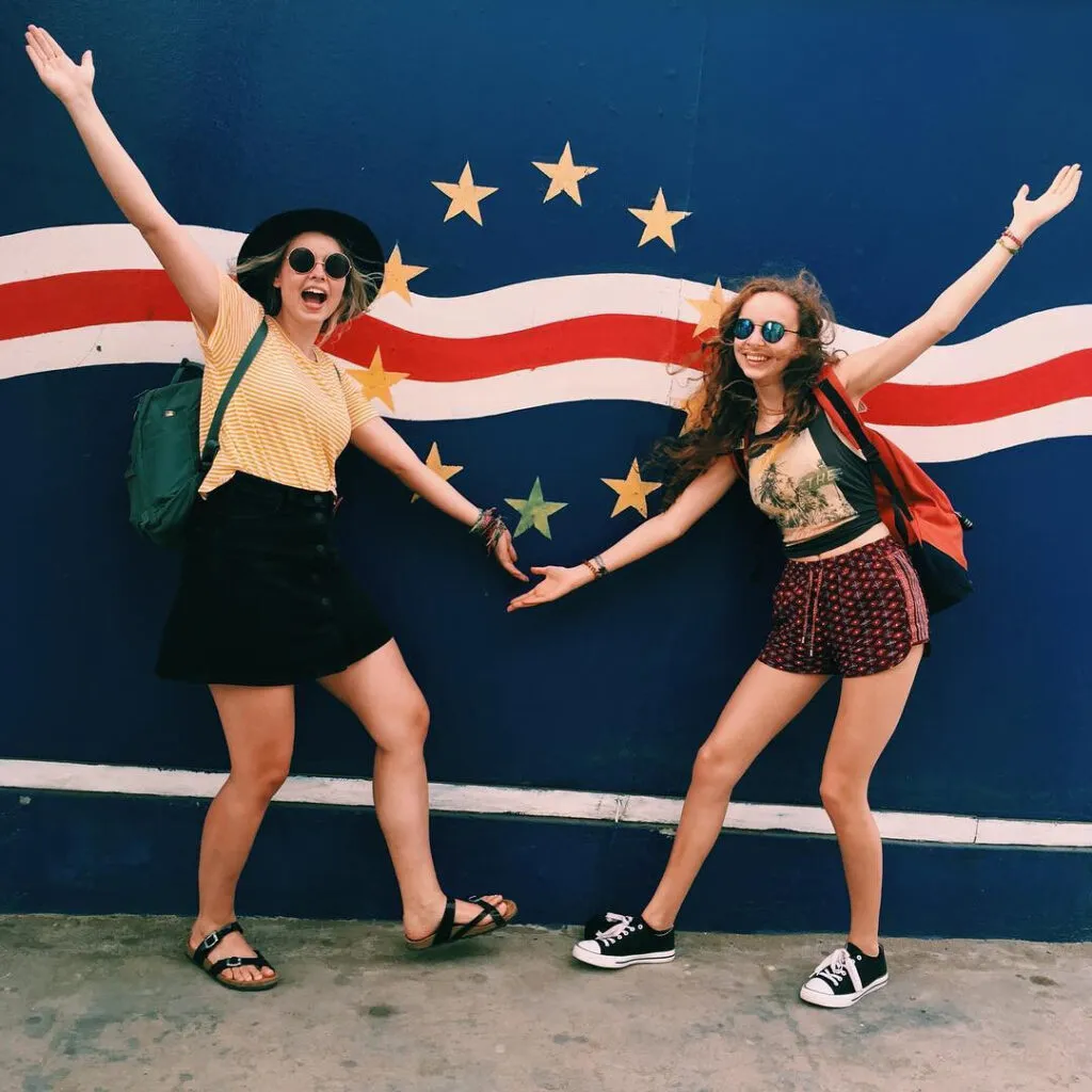 Twee jonge vrouwen poseren vrolijk met de armen in de lucht voor een grote, artistieke muurschildering van de Kaapverdische nationale vlag, op het eiland Sal.