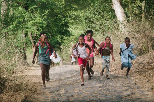 Een groep vrolijk lachende kinderen rennt met rugzakken over een onverharde weg door de natuur in het binnenland van Santiago, Kaapverdië.