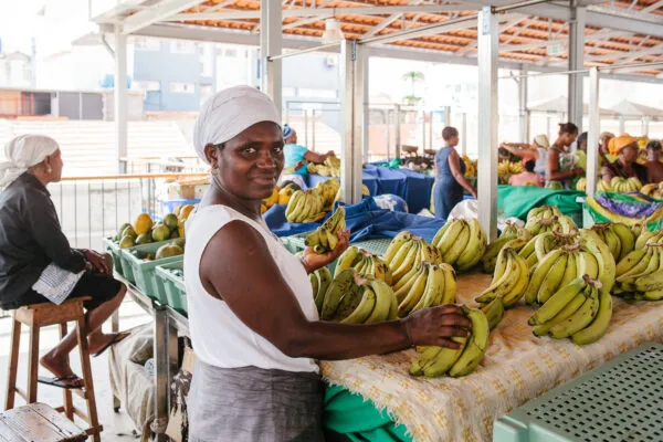 Lokale vrouw met hoofddoek staat achter een overdekte marktstal vol grote trossen groengele bananen op het eiland Santiago in Kaapverdië