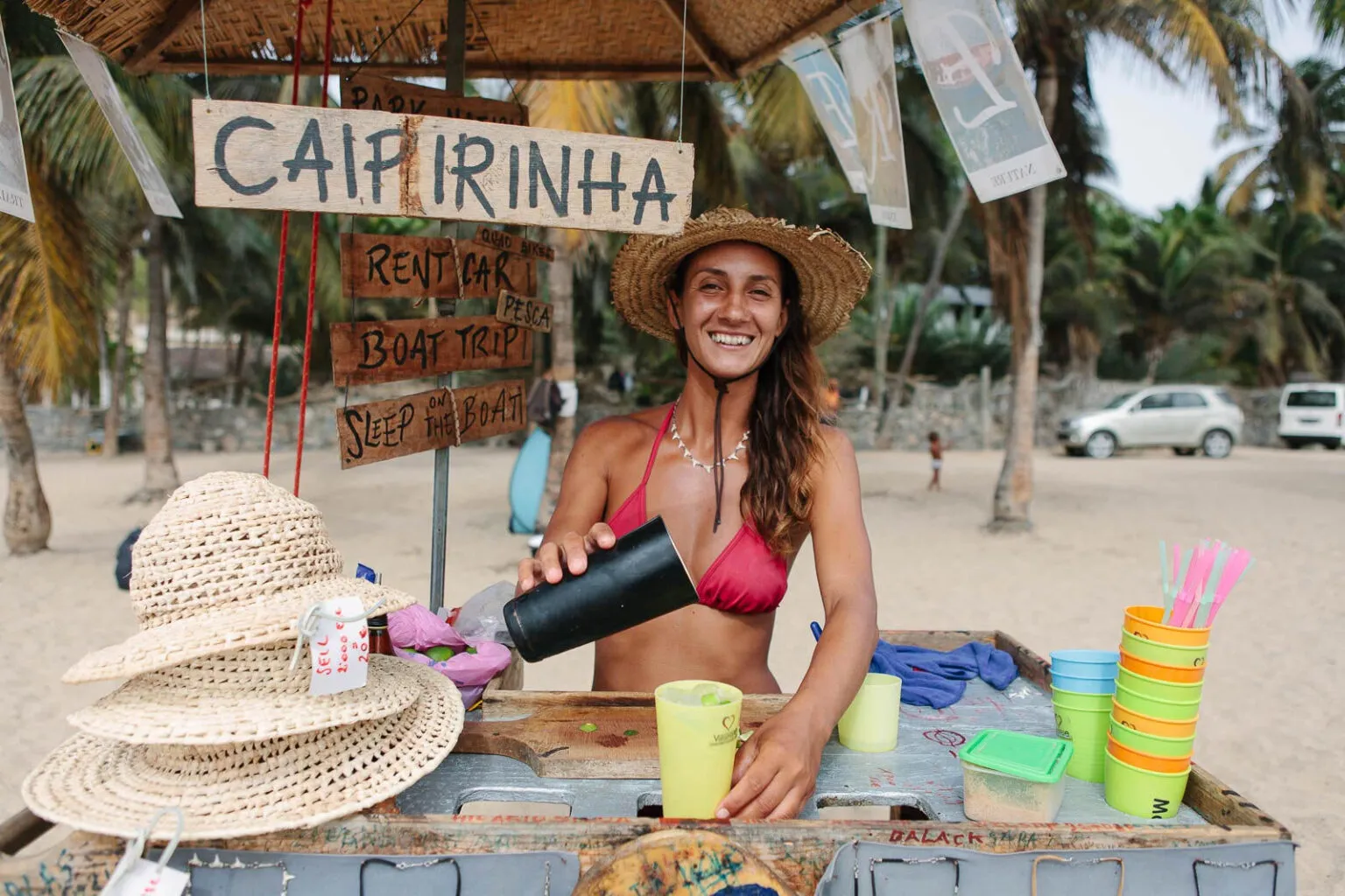 Lachende vrouw met een strooien hoed schenkt een drankje in, omgeven door palmbomen op het strand van Tarrafal op Santiago in Kaapverdië
