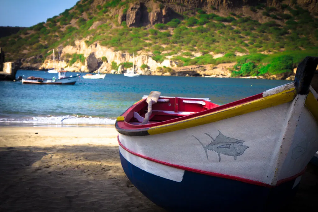 Een kleurrijke traditionele vissersboot ligt op het zandstrand van Tarrafal met een helderblauwe baai en groene bergen op de achtergrond op Santiago.