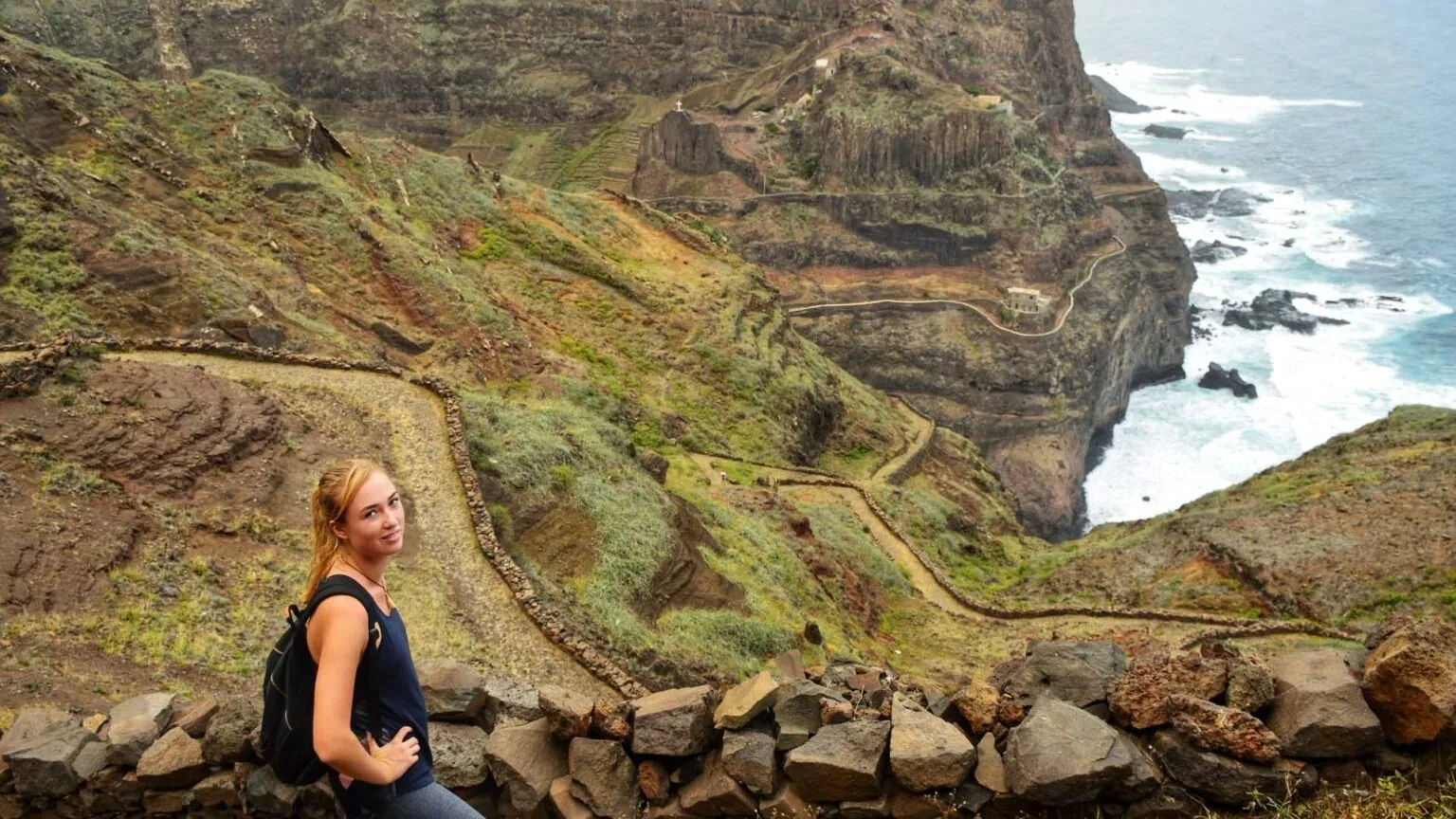 Reiziger Lina staat bij een stenen muurtje langs een kronkelend kustpad met op de achtergrond steile kliffen en de wilde oceaan op Santo Antão, Kaapverdië.