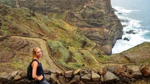 Reiziger Lina staat bij een stenen muurtje langs een kronkelend kustpad met op de achtergrond steile kliffen en de wilde oceaan op Santo Antão, Kaapverdië.